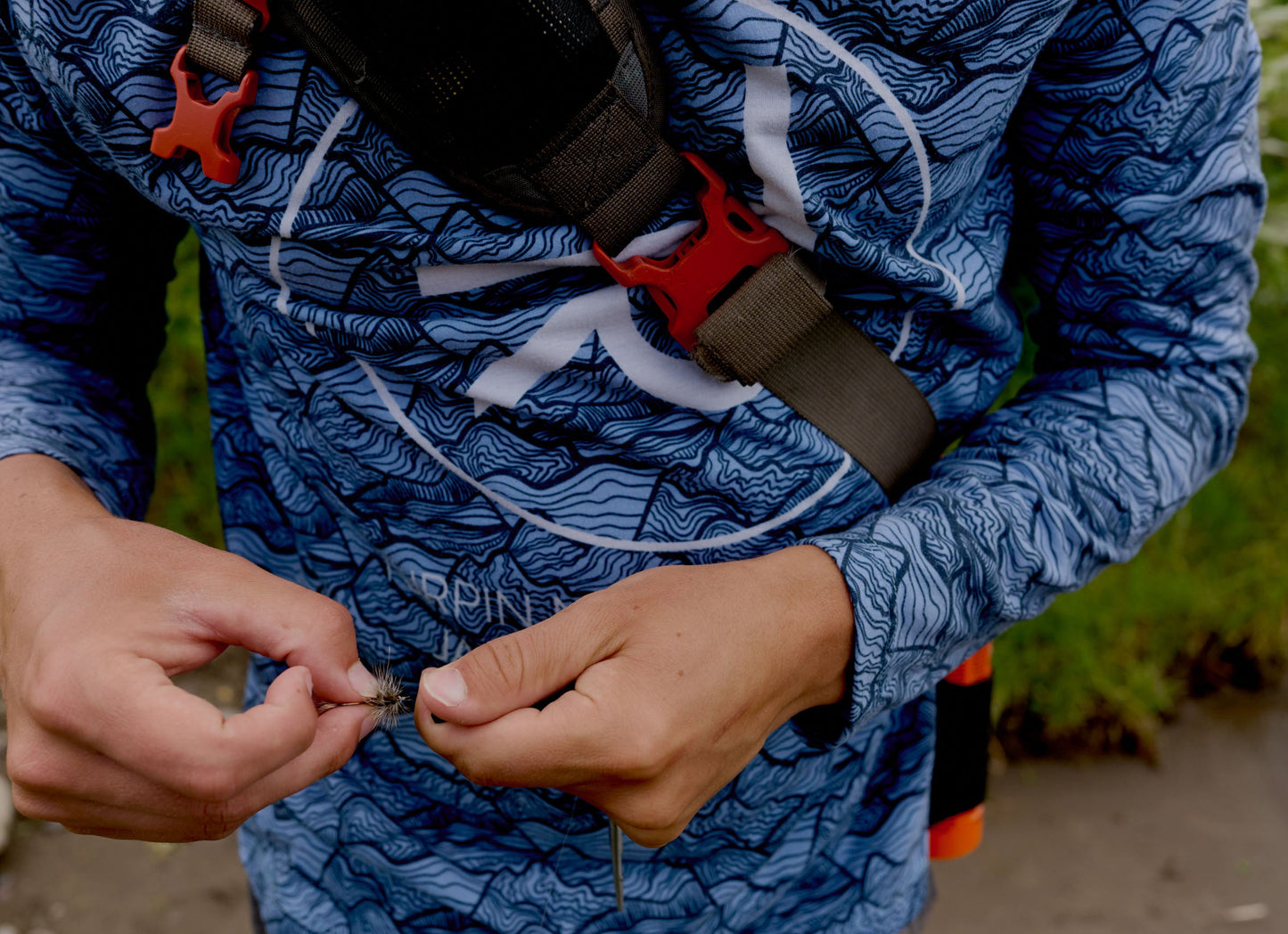 A close-up of a person outdoors wearing a blue long-sleeve patterned shirt with a black and red chest strap, while holding small fishing hooks in both hands.