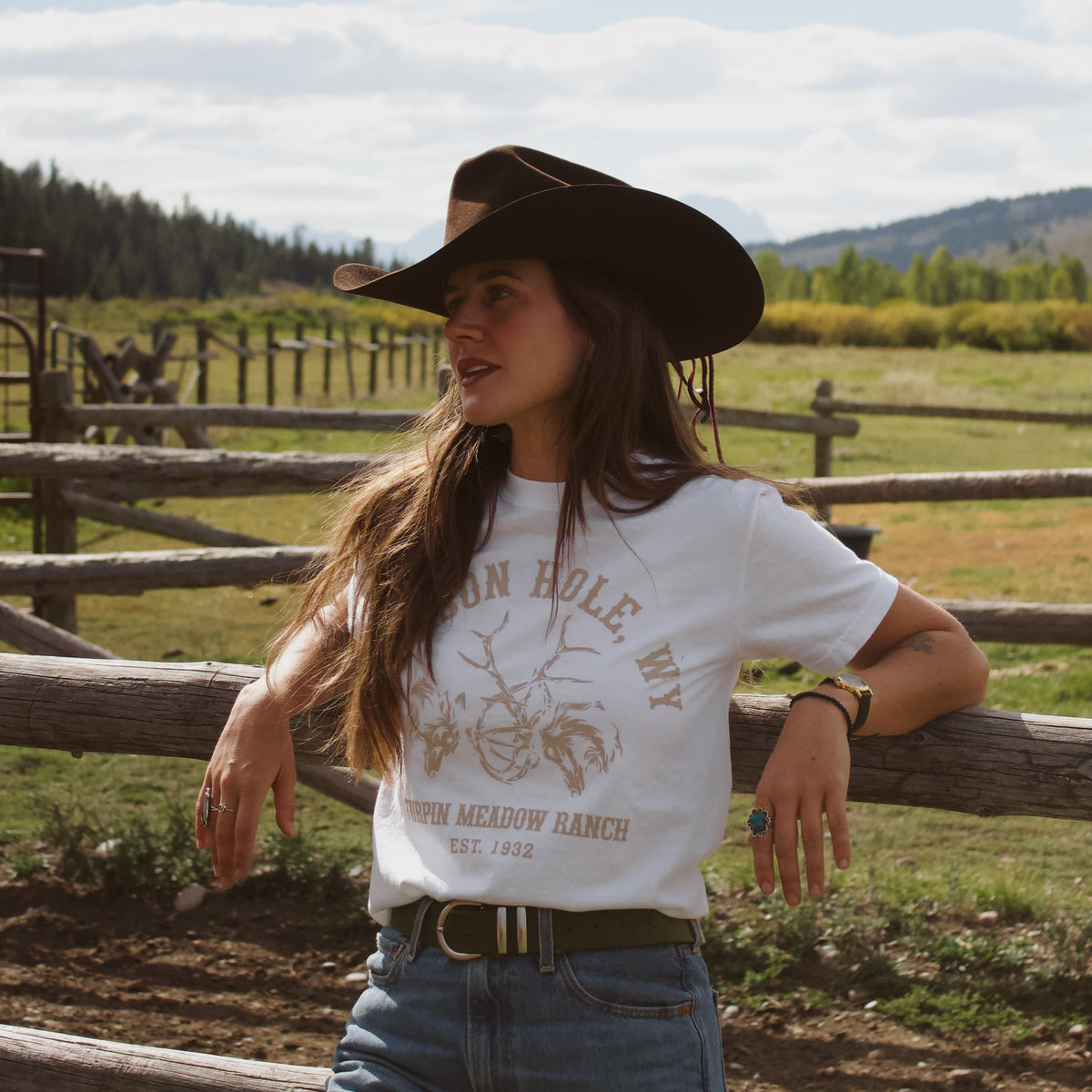 A woman wearing a white graphic t-shirt with a brown cowboy hat and blue jeans, standing outdoors in front of a wooden fence with a ranch and mountains in the background.